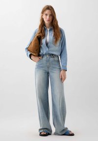 Vaaleansininen denim-paita, jossa on helmikitka, korkeavyötäröiset vaaleat farkut ja ruskea laukku. Sandaaleissa on sininen yksityiskohta.