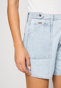 Mujer vistiendo unos pantalones cortos de mezclilla de tiro alto en azul claro con un botón y bolsillo, junto a una camiseta blanca metida por dentro, con la mano reposando cerca del muslo.