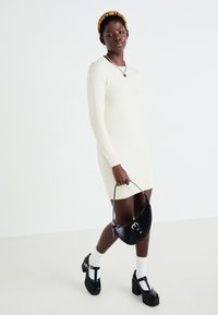 Robe blanche en tricot texturé avec manches longues, accompagnée d'un sac à main noir à motif crocodile et de chaussures à plateforme noires épaisses.