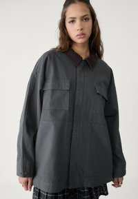 Übergroße graue Jacke mit braunem Kragen, ausgestattet mit zwei Fronttaschen und Knöpfen an den Manschetten. Hergestellt aus strapazierfähigem Stoff mit einer glatten Textur.
