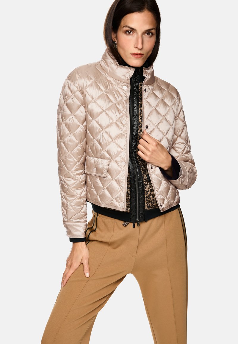 Gepolsterte beige Daunenjacke mit Stehkragen, Fronttasche und schwarzen Reißverschlüssen, über einem schwarzen Top mit Leopardenmuster getragen.