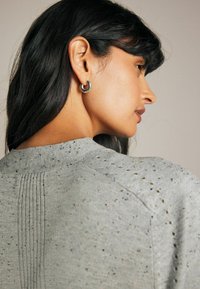 Grå sweater med tekstureret, spættet mønster, der har en ribbestrikket halsudskæring og bagdeller; sat sammen med sølvfarvede hoop øreringe.