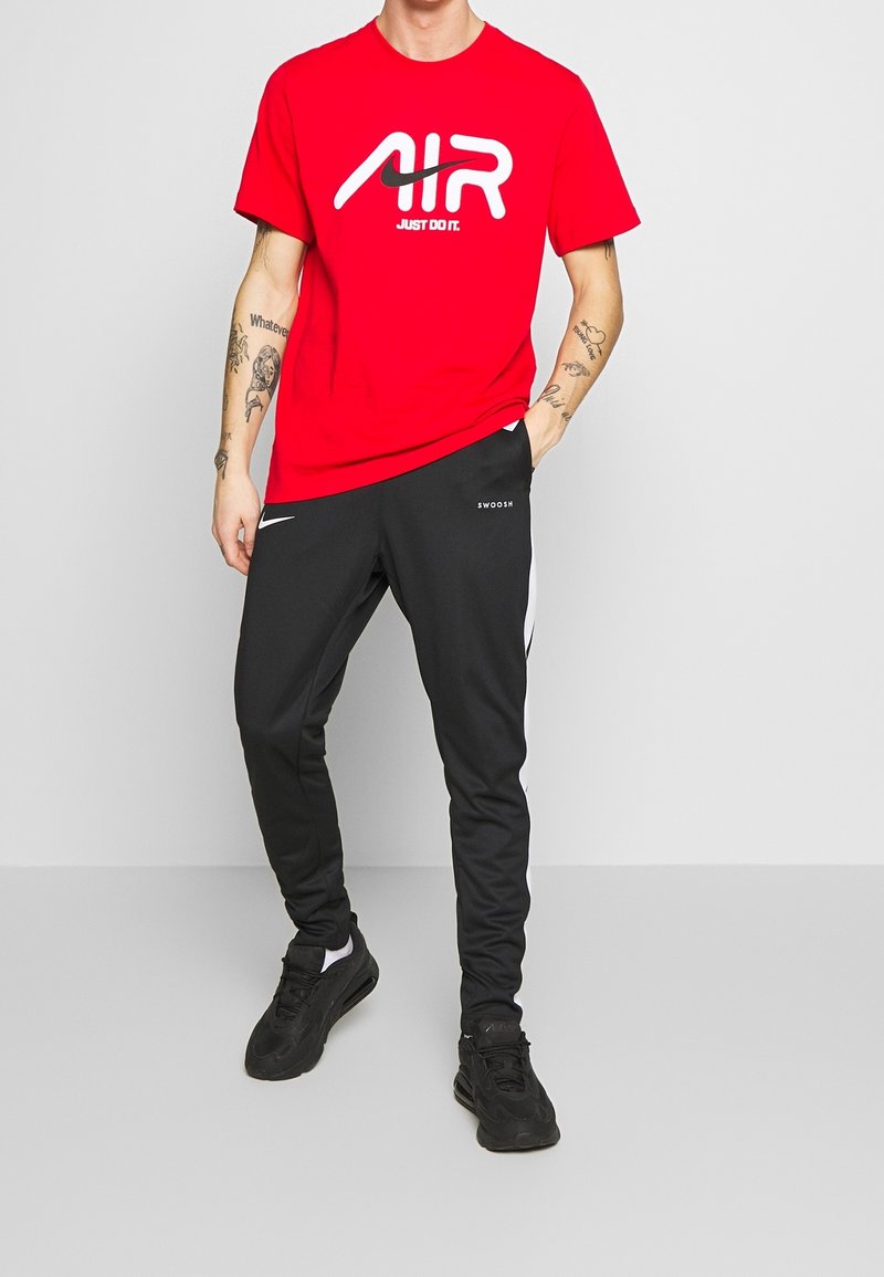 Punainen Nike t-paita, jossa valkoinen "AIR"-logo ja "JUST DO IT" -teksti, yhdistettynä mustiin urheiluhousuihin, joissa on valkoiset sivuraidat ja logo.