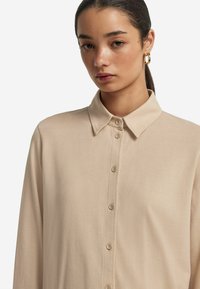 Beige långärmad skjorta med krage, med en knappslå och en slät, strukturerad tyg.
