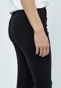 Svarta figurnära byxor i ett mjukt material, med en sidoficka och rak bendesign, visade från sidan med en hand vilande på låret.