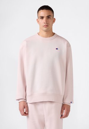 Hombre que lleva una sudadera Champion rosa claro y pantalones a juego, de pie sobre un fondo blanco liso.