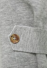 Gray knit textile featuring a textured finish, with a visible accent of a round, brown button on a tailored fabric strip.