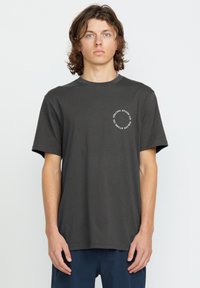 T-shirt en coton gris avec un col rond, des manches courtes et un accent de logo circulaire sur la poitrine. Texture douce, coupe décontractée, design minimal.