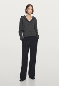 Dunkelgrauer Strickcardigan mit Kapuze, Knopfverschluss und gerippten Bündchen, kombiniert mit schwarzen gestreiften Hosen und schwarzen Schuhen.