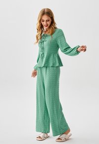 Top peplum verde chiaro testurizzato con fiocco frontale, abbinato a pantaloni a gamba larga nello stesso tessuto. Indossato con sandali bianchi a ciabatta.