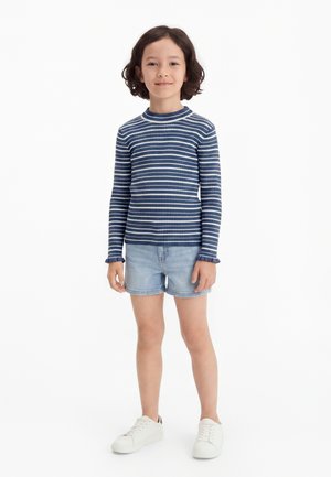 Barn med kort krøllet mørkt hår står iført en blå og hvidstribet langærmet top, lyse denimshorts og hvide sneakers på en hvid baggrund.