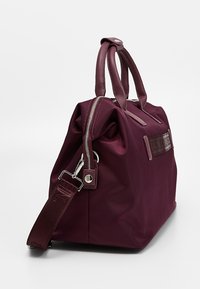 Viininpunainen nailonlaukku, jossa on kaksikahvaiset kahvat ja irrotettava olkahihna. Vetoketjullinen suljenta ja yksivärinen muotoilu. Koristeltu logoyksityiskohdalla.
