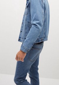 Chaqueta de denim azul con un corte clásico, que cuenta con un bolsillo frontal con parche y cierre de botones. Llevada sobre unos jeans azules a juego.