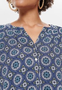 Blusa blu con motivo a cerchi in teal, bianco e verde. Presenta un collo a V e bottoni lungo la parte anteriore. Tessuto morbido.