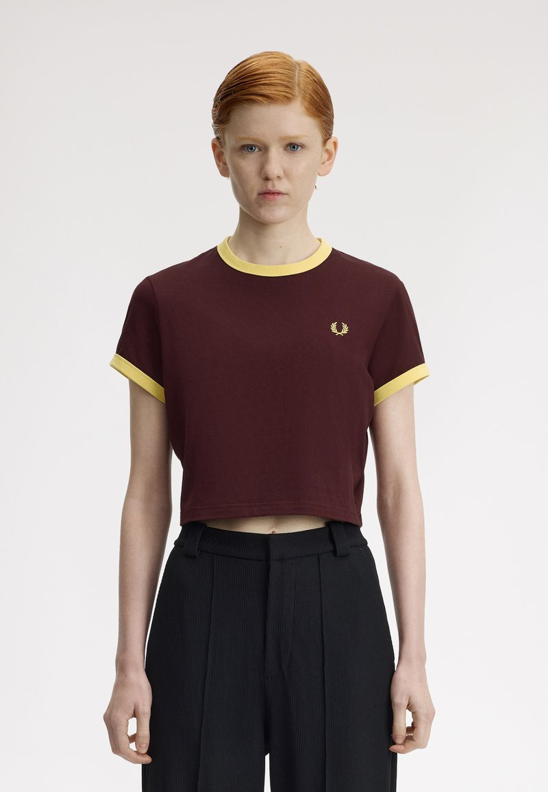 Jeune personne aux cheveux roux portant un t-shirt court bordeaux avec bordure jaune et un pantalon noir taille haute, debout devant un fond uni.