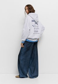 Felpa grigia con stampa grafica che presenta un cane e testo sul retro, abbinata a pantaloni in denim blu larghi. Stile casual, vestibilità rilassata.