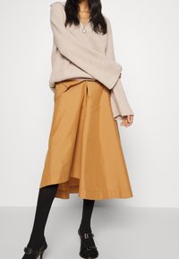 Beiger Rippstrickpullover mit Reißverschluss am Ausschnitt, kombiniert mit einem knieumspielenden, rostfarbenen Rock und schwarzen Strumpfhosen. Schwarze Sandalen mit Absatz setzen Akzente.