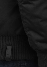 Black jacket with a textured fabric, featuring a ribbed hem and a leather tag labeled "SOLID." Close-up view emphasizes material and stitching.