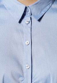 Chemise bleue claire à col avec une texture lisse, présentant une rangée de boutons blancs sur le devant. Remarquable pour son design simple.