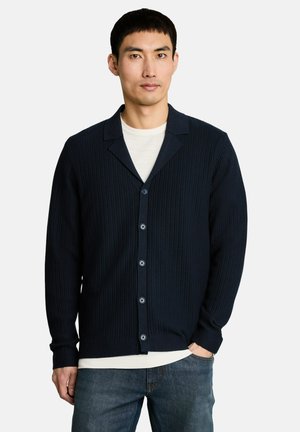 Mann trägt eine dunkelblaue gerippte Strickjacke über einem weißen Hemd mit blauen Jeans, steht vor einem schlichten hellen Hintergrund.