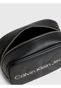 Borsetta in ecopelle nera con doppia chiusura a zip, con logo in rilievo "Calvin Klein Jeans" in oro. Texture liscia con design minimalista.