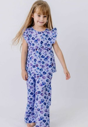 Young girl with long blonde hair wearing purple floral ruffled sleeveless top and matching pants, standing against plain background.