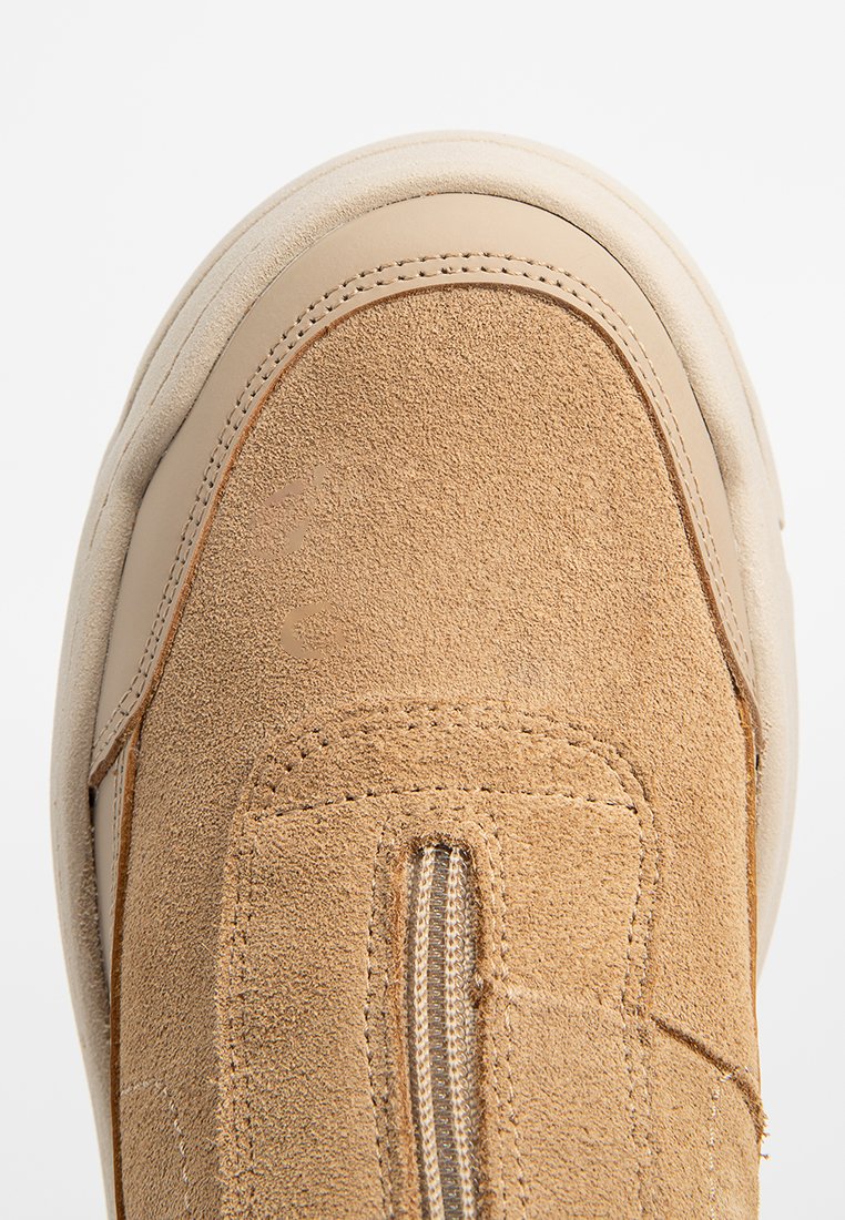 Zapatos de ante en tono marrón claro con punta redonda, que cuentan con un cierre de cremallera lateral y una suela de goma crema contrastante. Textura suave y mate con detalles de costura.