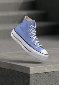 Μπλε ψηλό αθλητικό παπούτσι με λευκή σόλα και κορδόνια, αιωρούμενο στον αέρα πάνω από μια τσιμεντένια άκρη με σκάλες στο φόντο.