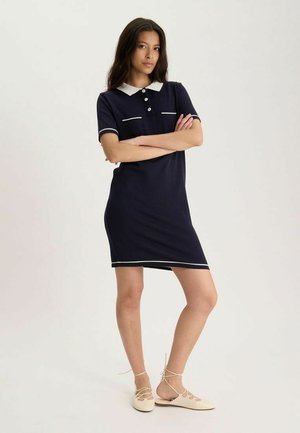 Jeune femme debout les bras croisés portant une robe polo bleu marine avec des bordures blanches et des chaussures plates beiges à lacets.