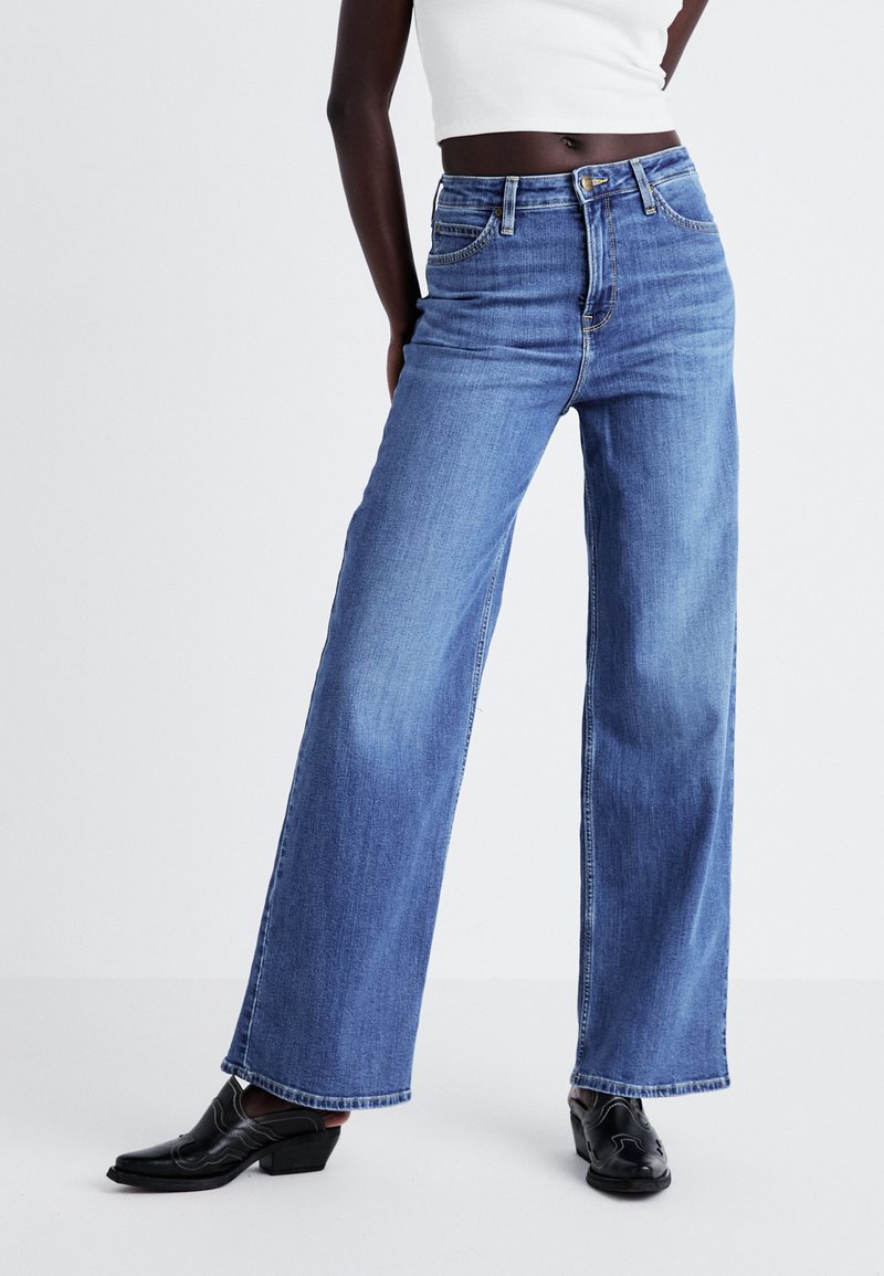 Model wearing high-waisted blue wide-leg jeans with five pockets and black heeled slip-on shoes, standing with one hand on hip.