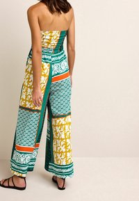Traje pantalón con un top sin tirantes de diseño floral en amarillo y blanco, pantalones de pierna ancha con patrones en verde, naranja y blanco, y sandalias negras.