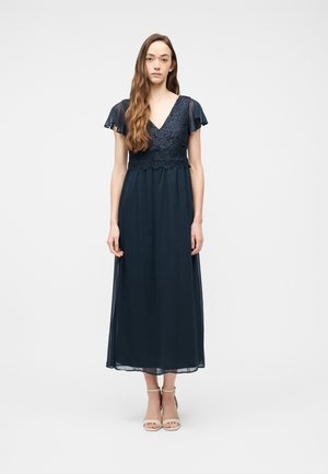 Mujer con cabello largo y castaño, vestida con un vestido de encaje azul marino con mangas cortas semitransparentes y tacones abiertos beige, de pie frente a un fondo liso.