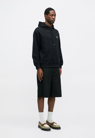 Sweat à capuche noir en tissu doux avec une poche avant, associé à un short noir de longueur genou et des chaussures bicolores distinctives.