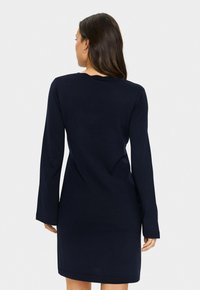 Donna con lunghi capelli scuri che indossa un vestito in maglia a maniche lunghe di colore blu navy, in piedi con la schiena rivolta verso un fondo chiaro e uniforme.