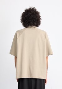 Persona con cabello rizado que lleva una camisa corta beige holgada y pantalones negros, vista desde atrás contra un fondo blanco simple.