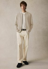 Hellbeige gestrickte Zip-Jacke über einem wei�ßen T-Shirt, kombiniert mit cremefarbenen Hosen und schwarzen Wildlederschuhen. Einfaches, maßgeschneidertes Design.