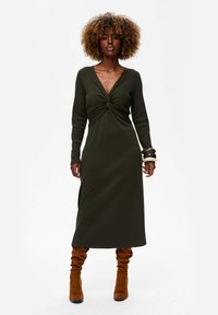 Długa oliwkowozielona sukienka z węzłem z przodu, długimi rękawami i slim fit, w połączeniu z brązowymi, wysokimi botkami o luźnym kroju.