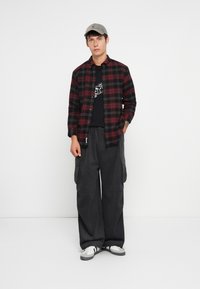 Joven con gorra gris, camisa de cuadros roja y negra sobre una camiseta gráfica negra, pantalones cargo oscuros anchos y zapatillas blancas, de pie contra una pared blanca.