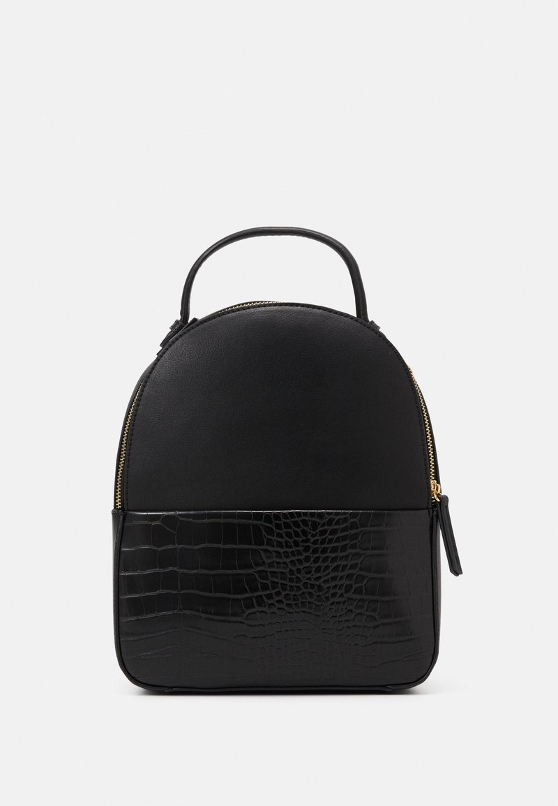 Sac à dos en cuir noir avec dessus lisse, bas en cuir à motif crocodile texturé, poignée supérieure unique et tirette de fermeture éclair en or sur fond blanc.