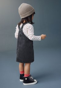 Fille portant une robe salopette en jean gris par-dessus une chemise blanche à manches longues avec des pois rouges, des baskets montantes noires et un bonnet en maille gris.