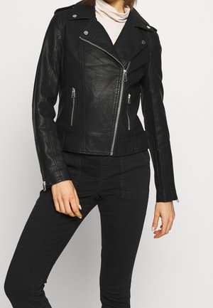 Vrouw draagt een zwarte nepleren motorjas met zilveren ritsen en zwarte broek, gecombineerd met een licht coltruitje.