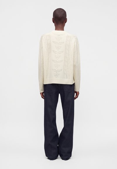 Personne aux cheveux courts portant un pull en laine crème avec un motif de feuilles et un pantalon large sombre, debout de dos sur un fond uni.