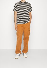 T-shirt à rayures noires et blanches à manches courtes avec une poche poitrine, associé à un pantalon cargo orange avec des poches latérales et des ourlets élastiques. Baskets blanches.