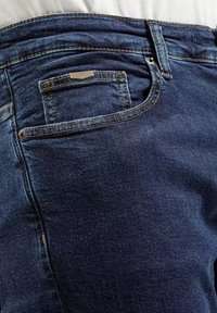 Ciemnoniebieskie dżinsy o klasycznym pięciokieszeniowym fasonie. Posiadają kontrastowe szwy, małą metalową tabliczkę z logo oraz gładką fakturę.