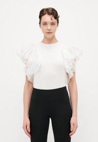 Balts tops ar caurspīdīgām, ruffled piedurknēm, pieguļošs dizains un apaļa kakla izgriezuma. Kombinēts ar melnām legingām. Minimālistiska un eleganta izskata.