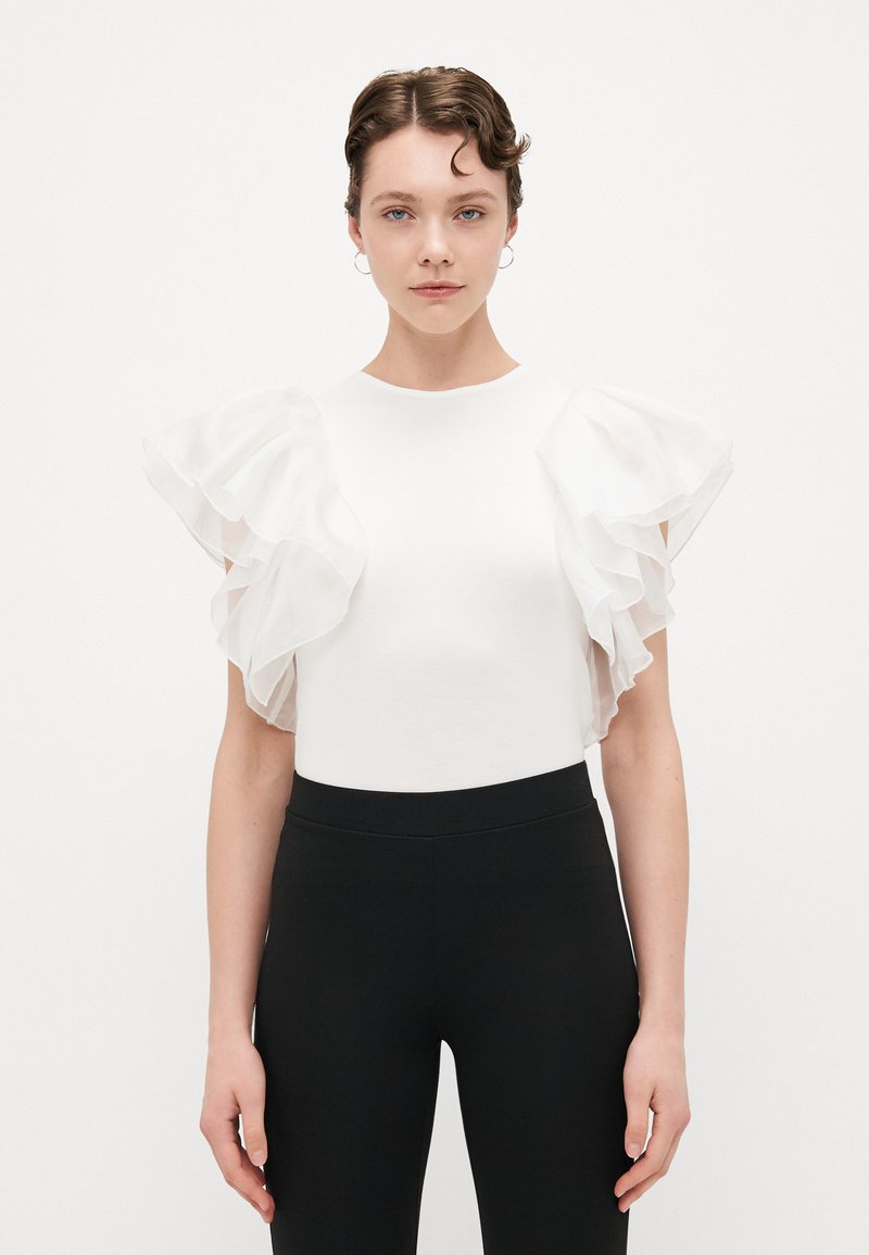 Balts tops ar caurspīdīgām, ruffled piedurknēm, pieguļošs dizains un apaļa kakla izgriezuma. Kombinēts ar melnām legingām. Minimālistiska un eleganta izskata.