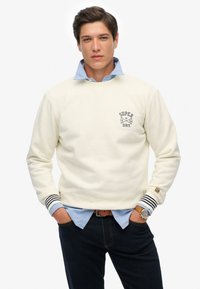 Sudadera crema con puños acanalados a rayas, con un pequeño logo en el pecho. Llevada sobre una camisa de collar azul claro. Jeans oscuros.