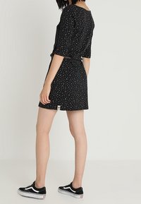 Svart klänning med färgglada prickar, korta ärmar, midjeband och en etikett vid kanten. Bärs med svarta sneakers.