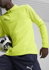 Felgeel sportshirt met een kwart-rits, gestructureerde stof en PUMA-logo. Model houdt een voetbal vast, draagt een marineblauwe short.