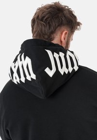 Svart hettegenser med stor, brodert hvit tekst på hetten. Har en avslappet passform og myk tekstur. Ingen synlig utstyr eller detaljer.